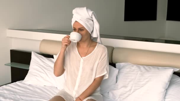 穿着浴衣和毛巾的女孩坐在床上喝咖啡。4k，慢动作 — 图库视频影像