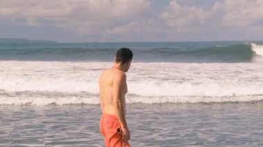 Turuncu şortlu çıplak gövdesi olan genç atletik, bronzlaşmış, bin yıllık bir adam güneşli bir günde dalgalar ve köpüklerin ortasında Hint Okyanusu 'nda duruyor. Turist tatilde. Tropikal Ada. 4k, ağır çekim.