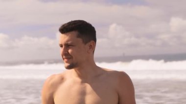 Güneşli bir günde kızgın dalgalar ve köpüklerin arasında çıplak gövdesiyle Hint Okyanusu 'nda duran genç, bronz tenli, bin yıllık bir adamın portresi. Tatilde bir turist. Tropikal ada. 4k, ağır çekim.