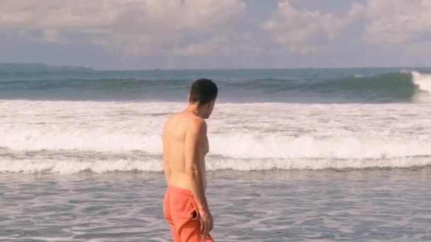Jovem atlético, bronzeado, homem milenar com um tronco nu em shorts laranja fica no Oceano Índico em meio a ondas furiosas e espuma em um dia ensolarado. Turista de férias. Ilha Tropical. 4k, câmera lenta — Vídeo de Stock