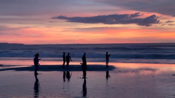 Napnyugtakor az emberek sziluettjei az óceán hátterében a parton. Festői, színes naplemente az Indiai-óceánon. Bali, Indonézia, 2022. április 1. — Stock videók