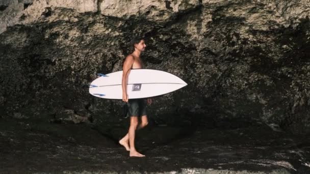Surfer. Bärtiger männlicher Millennial mit nacktem Oberkörper läuft mit Surfbrett unter der Achselhöhle an Felsen entlang. Zeitlupe, 4k. Indonesien, Bali, 13. März 2022 — Stockvideo