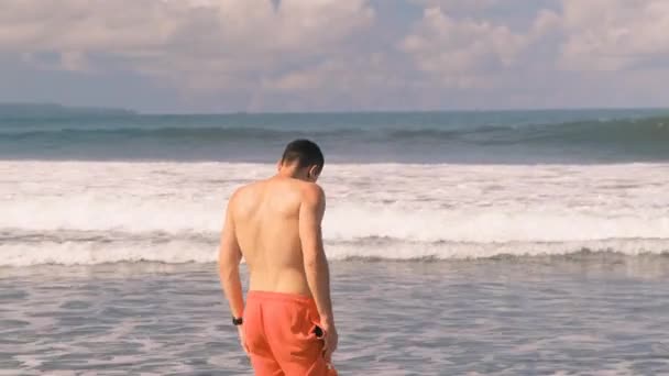 Jovem atlético, bronzeado, homem milenar com um tronco nu em shorts laranja fica no Oceano Índico em meio a ondas furiosas e espuma em um dia ensolarado. Turista de férias. Ilha tropical. — Vídeo de Stock