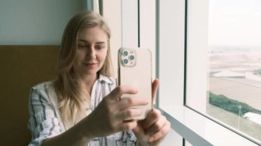 Blonde girl takes a selfie sitting next to the window smiling looking at the smartphone. Happy beautiful woman takes pictures of herself on a mobile phone wearing a shirt. Slow motion, 4k