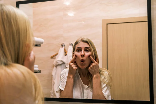 Second chin, obesity. Portrait of 30 year old girl lifting her cheeks up using her fingers while looking at mirror in bathroom. Aging and appearance problems.. Facelift, plastic surgery, facebuilding