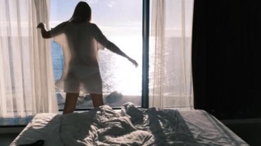 Şeffaf bornozlu bir kadın güneşli bir günde denize bakan panaramik bir pencerenin zemininde dans eder. Mutluluk, neşe ve tatmin duygusu. Arka plan.