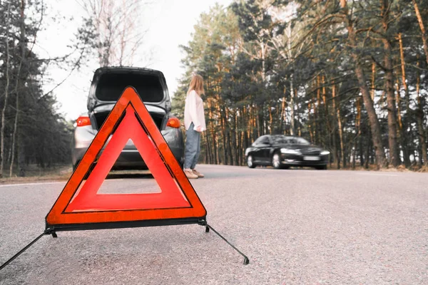 Red emergency triangle on the road with a blurry car in the background and a desperate woman catching passing cars waiting for help. Focus on emergency sign. Accident and broken car on the road.