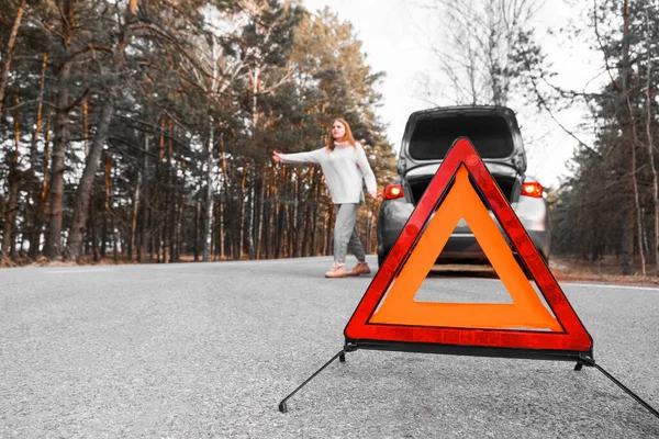 Red emergency triangle on the road with a blurry car in the background and a desperate woman catching passing cars waiting for help. Focus on emergency sign. Accident and broken car on the road.