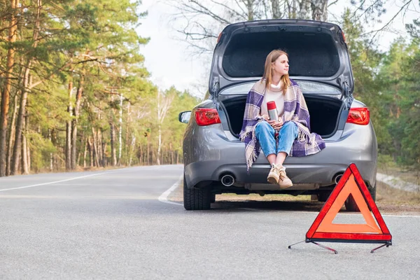 Red emergency triangle with car and woman sitting in the trunk with thermos in the background waiting help. Broken car on the road.