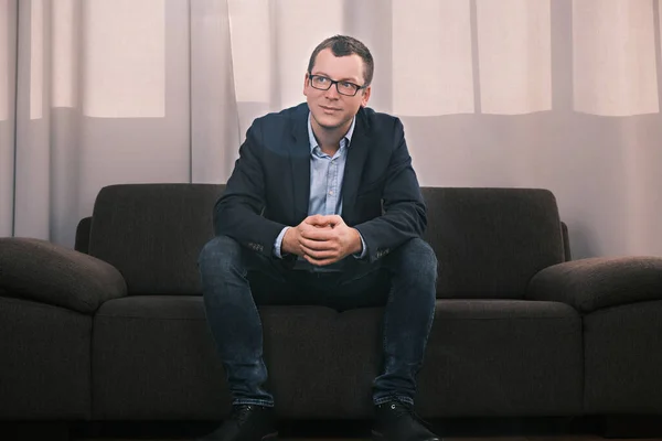 Portrait of caucasian man wearing jacket and glasses sitting on sofa in closed position with folded hands looking to the side against of alarge window with curtain. Success and business concept.