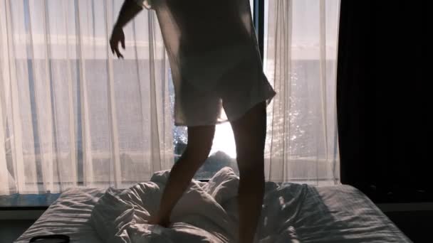 在阳光灿烂的日子里 穿着透明浴衣的女人在一个全景窗的背景下在床上跳舞 可以俯瞰大海 快乐和满足的情感 背景色 — 图库视频影像