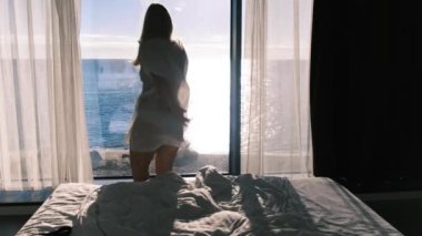 Şeffaf bornozlu bir kadın güneşli bir günde denize bakan panaramik bir pencerenin zemininde dans eder. Mutluluk, neşe ve tatmin duygusu. Arka plan.