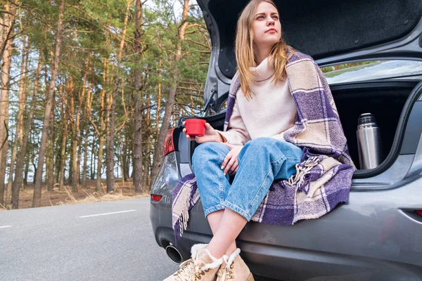 Young woman sitting on car trunk with coffee cups and thermos on the side of the road in the forest in warm fall or spring day. Time for yourself. Contemplation and rest in solitude