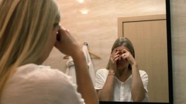 Yaşlanma. Gevşek deri. Banyodaki aynaya bakarken yüz gerdirme egzersizi yapan genç bir kadın. Facebook binası. Modern bakım, yüz kaslarının gevşemesi. Yüz gerdirme, estetik ameliyat