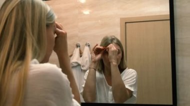 Yaşlanma. Gevşek deri. Banyodaki aynaya bakarken yüz gerdirme egzersizi yapan genç bir kadın. Facebook binası. Modern bakım, yüz kaslarının gevşemesi. Yüz gerdirme, estetik ameliyat