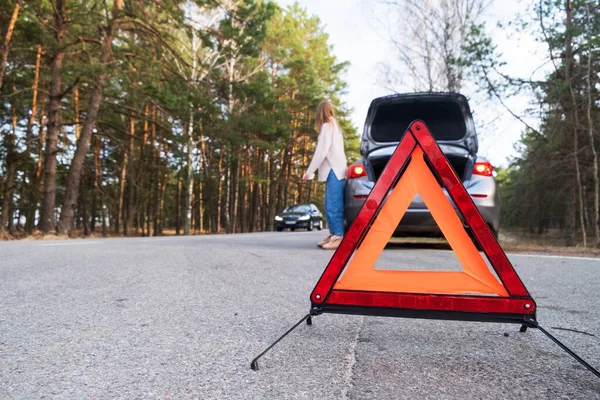 Red emergency triangle on the road amid a blurry car and a woman in despair catching passing cars waiting for help. Focus on the emergency sign. Accident and broken car on the road