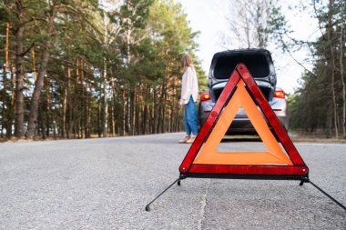 Red emergency triangle on the road amid a blurry car and a woman in despair catching passing cars waiting for help. Focus on the emergency sign. Accident and broken car on the road