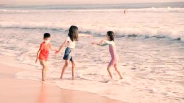 Asian children indulge and play in the water. Little girls and a boy are waiting for the waves, standing in the ocean and enjoying their childhood at sunset. Bali, Indonesia, June 28, 2022