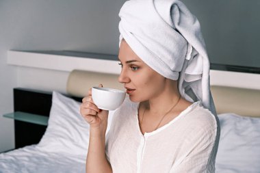 Side view portrait of a young millennial woman drinking morning coffee or tea while sitting on the bed in the bedroom with a white towel on her head in a bathrobe after a shower. Morning concept