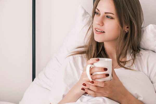 Young attractive woman lies in bed after waking up with white cup in her hand looking out the window. Morning routine, time for yourself. contemplation and enjoyment. Psychological self care.