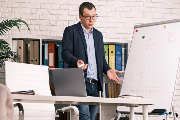 Businessman is giving an online presentation while having a video conference. Serious man in glasses and a jacket shows a development chart on a white board while in his office or home office.