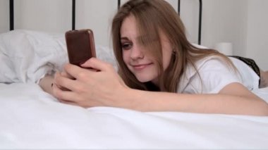 Young happy smiling woman using phone while lying in bed in the morning. Awakening. Girl reads good news, social networks, online communication. morning routine