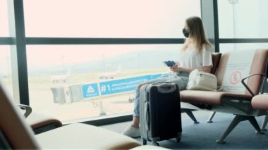 Woman passenger is waiting for her flight while sitting at airport against large panoramic window with suitcase next to her. Girl looks at her passport and ticket, check the gate. Travel, immigration.