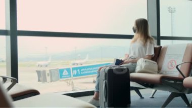 Woman passenger is waiting for her flight while sitting at airport against large panoramic window with suitcase next to her. Girl looks at her passport and ticket, check the gate. Travel, immigration.
