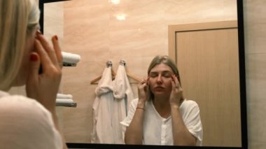 Yaşlanma. Gevşek deri. Banyodaki aynaya bakarken yüz gerdirme egzersizi yapan genç bir kadın. Facebook binası. Modern bakım, yüz kaslarının gevşemesi. Yüz gerdirme, estetik ameliyat