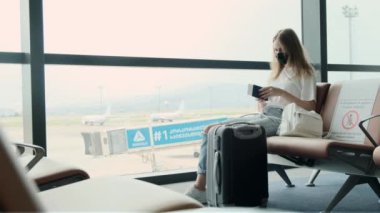 Woman passenger is waiting for her flight while sitting at airport against large panoramic window with suitcase next to her. Girl looks at her passport and ticket, check the gate. Travel, immigration.