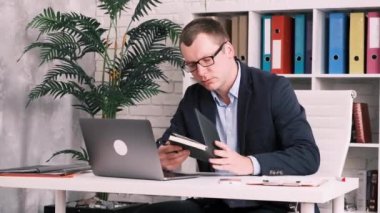 Office worker, businessman or businesswoman in glasses and a suit works with a notepad and laptop while sitting in office at the desk against backdrop of shelves with folders. White-collar worker