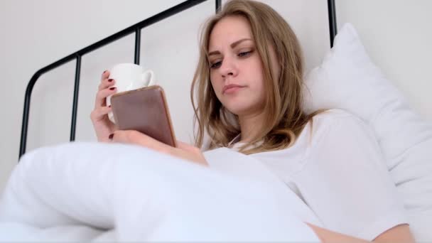 Serious Concentrated Woman Uses Phone While Lying Bed Morning Cup — Vídeo de Stock