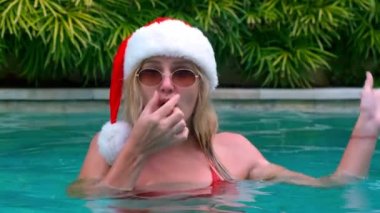 Güneş gözlüklü genç bir kadının Noel portresi, Noel Baba şapkası ve kırmızı bir mayo kameraya bakarken havuza dalıyor. Genç komik kadın tropiklerde Noel 'i kutluyor.