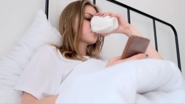 Ciddi, konsantre bir kadın sabahları elinde fincanla yatıp kahve içerken telefonu kullanır. Uyanış. Kötü haber okuyan kızlar, sosyal ağlar, online iletişim. Sabah rutini