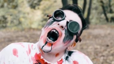Ağzından kan ve siyah sıvı çıkan zombi yüzünün yakın plan portresi. Makyajlı, havacılık gözlüklü, cam kırıkları olan bir adam kameraya ve suratını buruşturanlara bakıyor.