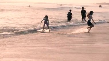 Asyalı çocuklar okyanusta oynuyorlar. Çocuklar gün batımında dalgalara atlayıp sahilde koşar ve çocukluklarının tadını çıkarırlar. Bali, Endonezya, 28 Haziran 2022