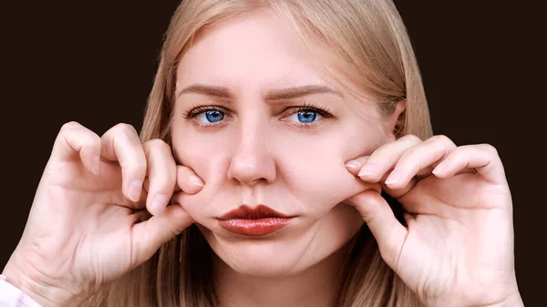 Second chin, obesity. Portrait of a 30-year-old woman pulling back the skin on her cheeks looks into the camera with a frustrated look. Problems of aging and appearance. Facelift, plastic surgery.