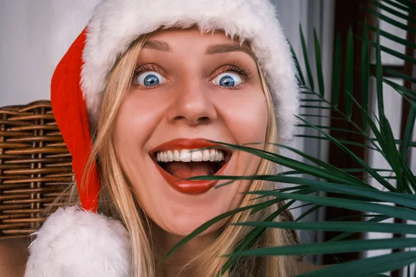Funny broad smiling blonde woman wearing santa hat looking at camera with surprised eyes. Girl celebrating Christmas in tropics. Festive mood. Christmas or New Year discounts