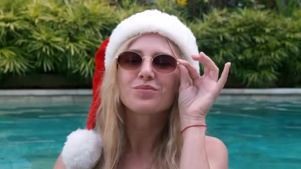 戴着桑塔帽和太阳镜的金发美女在游泳池里面对着绿色热带植物时看着相机 她走到游泳池边 用相机调情 热带岛屿的圣诞概念 — 图库视频影像