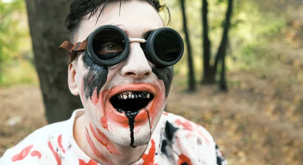 Portrait is close-up of face of zombie with traces of blood and black fluid flowing from the mouth. Man with makeup with aviation glasses with broken glass looks into camera and grimaces