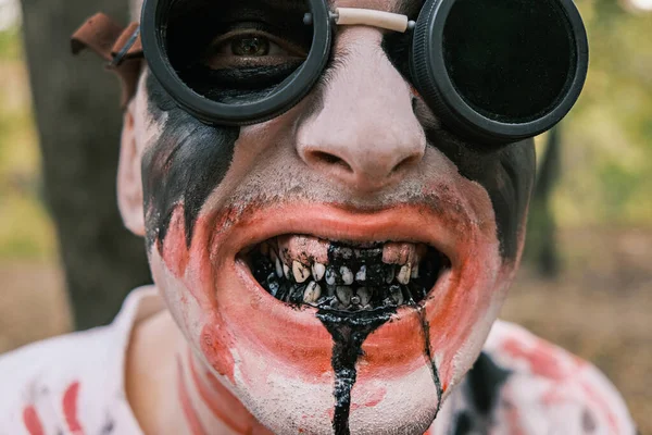 Close-up portrait of face of zombie with traces of blood and black fluid flowing from the mouth. Man with makeup with aviation glasses with broken glass looks into camera and grimaces