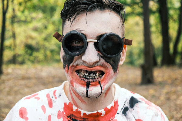 Portrait is close-up of face of zombie with traces of blood and black fluid flowing from the mouth. Man with makeup with aviation glasses with broken glass looks into camera and grimaces