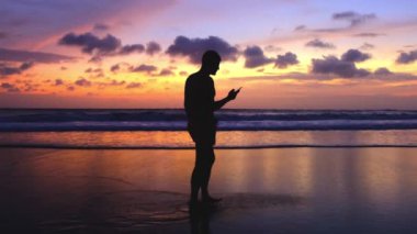 Sahilde yürüyen bir adamın silueti kızgın renkli günbatımına karşı, telefona bakıyor. Güzel bir manzaranın ve bir insanın yavaş çekim 4K görüntüsü. Adam okyanus boyunca yürürken cep telefonu kullanıyor.