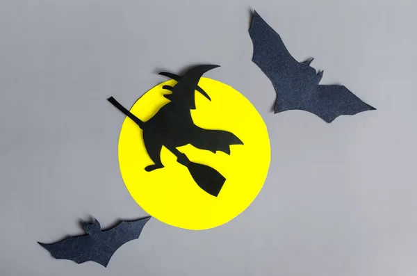 The shadow of a black witch in close-up flying on a broom surrounded by bats against a yellow moon.