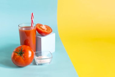 Şeffaf bir bardaktaki domates suyu reklamı ve domates dekoru, mavi bir podyum ve sarı-mavi arka plandaki tuz boşluğu kopyalar.
