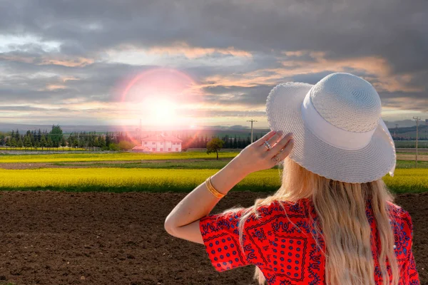 后视镜上的姑娘头戴白色的帽子 平静地看着黄色的油菜花田和乡间别墅上空的落日 — 图库照片