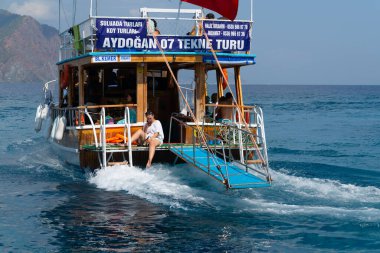 Adrasan, Antalya / Türkiye-25 Eylül 2020: Turist taşıyan bir tur teknesi denizde seyahat ediyor ve teknenin arkasındaki bir turist kadın cep telefonuna bakıyor.