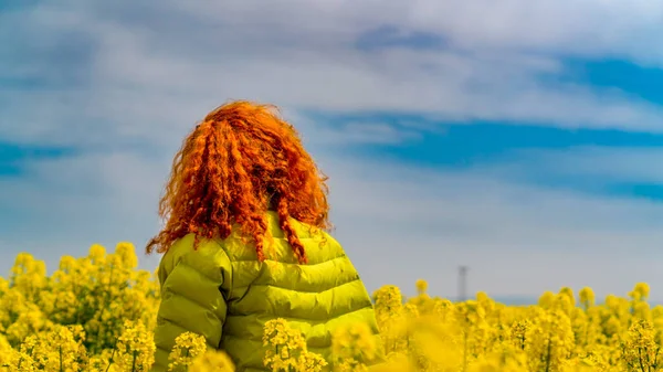 Back view of red hair woman in yellow coat in canola field