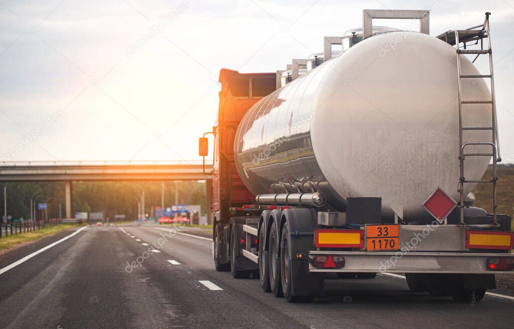 A truck with a tank trailer transports a liquid dangerous cargo on a highway against the backdrop of a sunset.