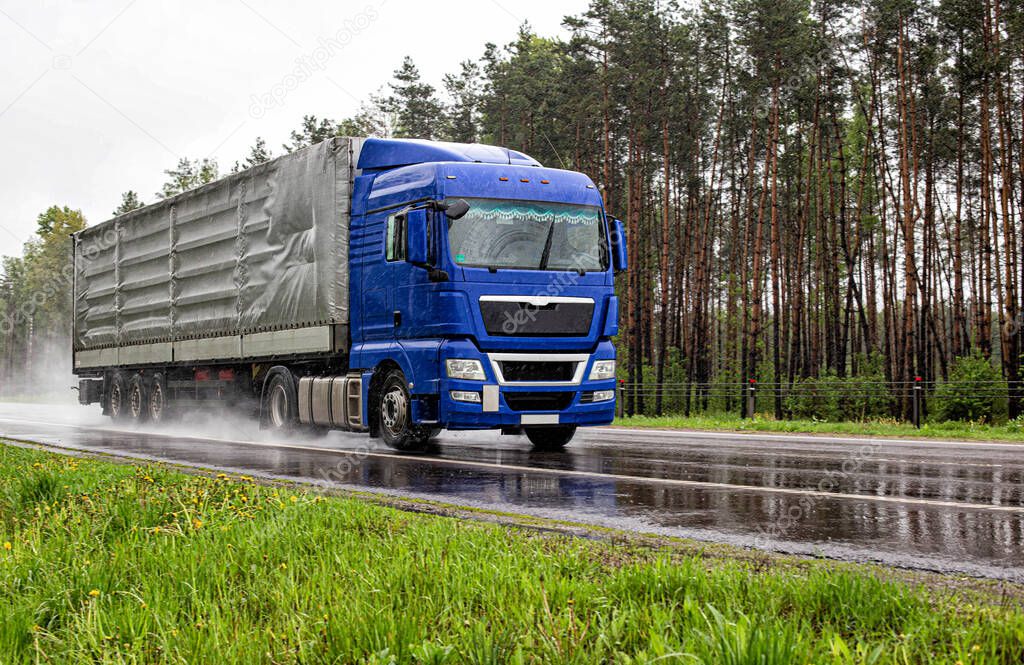 A truck with a tilt semi-trailer drives on the road in the rain and poor visibility in the summer against the backdrop of the forest. Cargo transportation and logistics concept